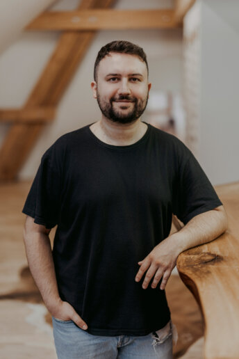 Mann in schwarzem T-Shirt lehnt entspannt an einem Holzgeländer in einem hellen Raum mit sichtbaren Dachbalken.